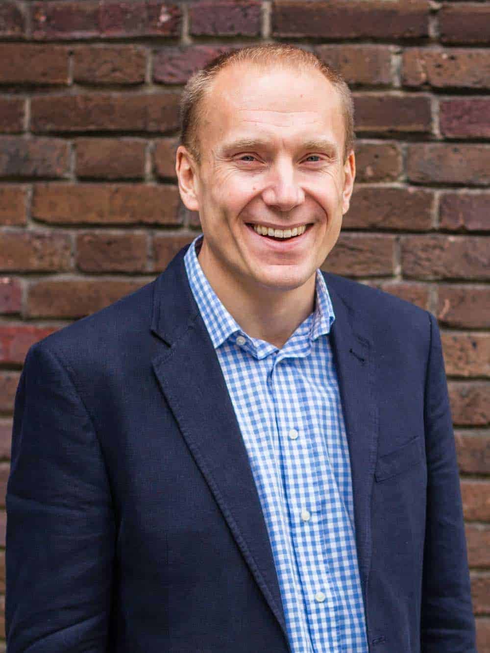 Smiling man in a navy blazer and blue check shirt standing against a brick wall.